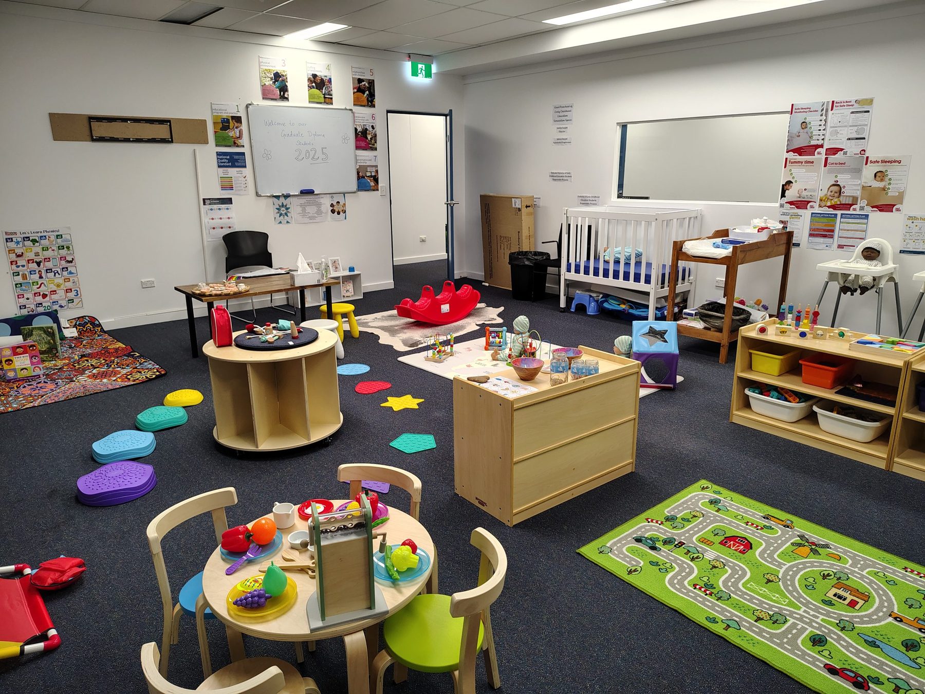 Play and activity area of the simulation room, with child-sized furniture, a crib and a welcome board for graduate diploma students